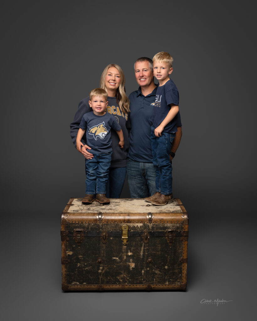 Jillian Duffey and her family (husband and 2 sons) pose for family portraits. They wear Montana State Bobcat gear and stand in front of a dark gray background. Her sons stand on a wooden trunk.