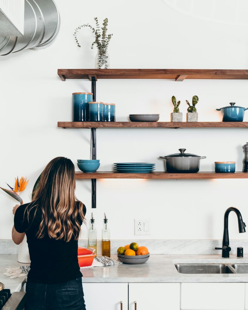 A woman sets flowers into a vase in her well-organized kitchen. The shelves are open, neat and clean and hold plates, cannisters and baking pots.