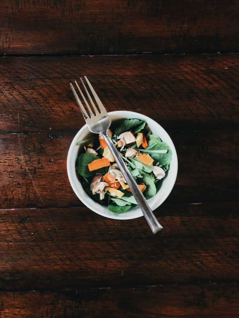 The view from over the top of a bowl of a fresh-looking salad with greens, nuts and veggies. The bowl sits on a dark wooden table and a fork rests across it.